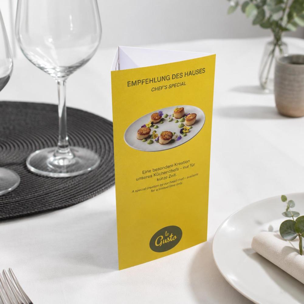 Bedruckter Prisma-Tischaufsteller mit gelber Gestaltung und Menüempfehlung auf einem gedeckten Restauranttisch.
